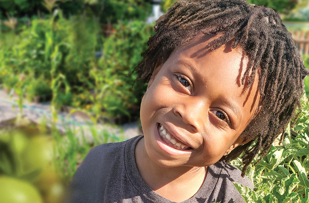 A little boy smiles, surrounded by green growing produce in a garden